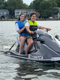 Two smiling people in blue and neon-green life jackets riding a gray Yamaha jet ski on a calm lake with waterfront homes and trees in the background.