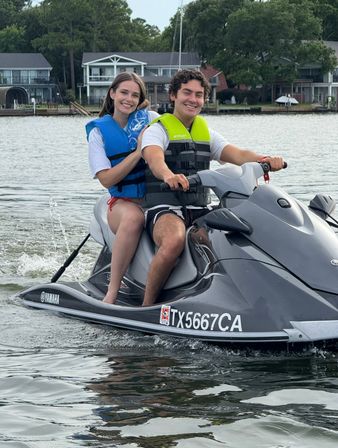 Two smiling people in blue and neon-green life jackets riding a gray Yamaha jet ski on a calm lake with waterfront homes and trees in the background.