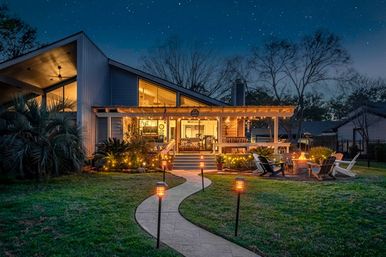 Cozy modern home backyard at night with a winding illuminated pathway leading to a covered patio decorated with string lights, outdoor seating around a fire pit, and a starry sky.