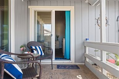 Inviting covered porch with two wicker chairs topped by blue-and-white striped pillows, a small round table with coffee and flowers, sliding glass door with turquoise curtain, gray vertical siding and a 'Welcome to Lake House' doormat.