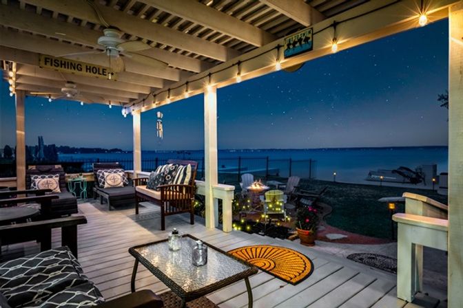 Cozy covered lakefront patio at night with string lights, cushioned seating, glass-top table, small fire pit, potted plants, and a dock across calm water under a starry sky.