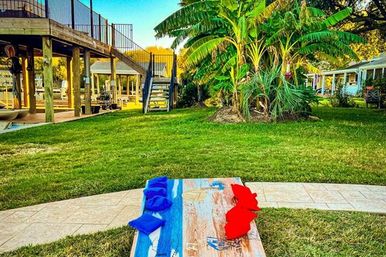 Cornhole board with red and blue bean bags on a patio edge in a tropical backyard, lush green lawn, banana palms and a raised wooden dock with stairs by the water.