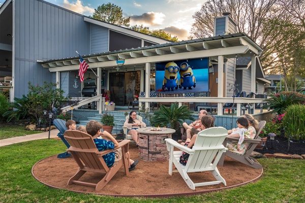 Backyard outdoor movie night at a suburban home with adults and children in Adirondack chairs around a stone fire pit, watching a large porch screen showing Minions under an evening sky.