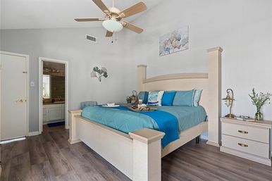 Bright master bedroom with light-wood king bed and teal blue bedding, wooden ceiling fan, nightstand with lamp and vase, ensuite bathroom doorway, and gray wood-look flooring