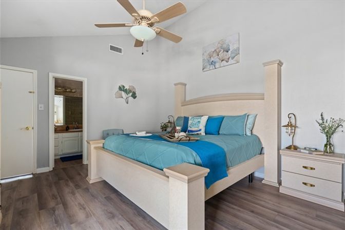 Bright master bedroom with light-wood king bed and teal blue bedding, wooden ceiling fan, nightstand with lamp and vase, ensuite bathroom doorway, and gray wood-look flooring