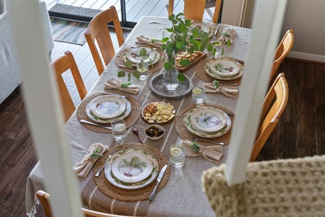 Inviting coastal-inspired dining table set for six with woven placemats, palm-print plates, linen napkins, glass water glasses with lemon, cheese and olive snacks, and a vase of greenery on a wooden table with natural wood chairs in a bright home dining room.