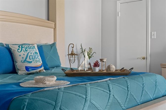 Lake-house bedroom with teal quilt, "Relax you're on Lake Time" pillow, straw hat and nautical tray holding a sailboat model and succulents.