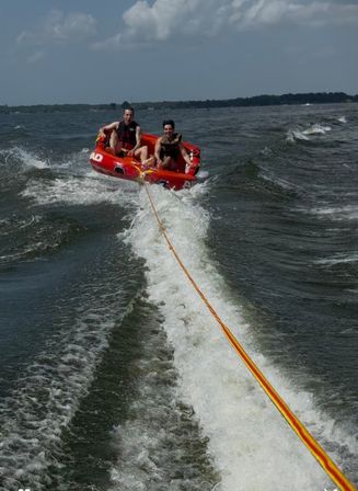 Two people in life jackets riding a red inflatable towable tube across a lake, held by a yellow-orange tow rope behind a speedboat’s foamy wake under a partly cloudy sky.