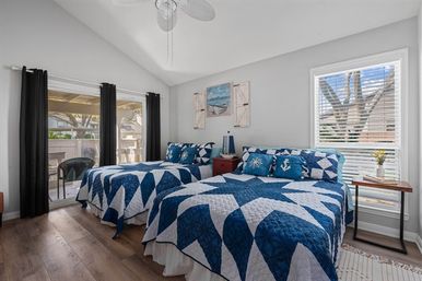 Bright coastal-style bedroom with two beds in matching blue star quilts and nautical pillows, hardwood floors, vaulted ceiling fan, sliding glass door to a patio and a sunlit window with white blinds.