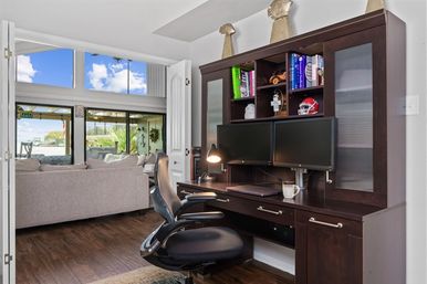 Bright home office with dark wood desk and dual monitors, ergonomic chair and bookshelves, opening to a living room and sliding doors with a waterfront patio view.