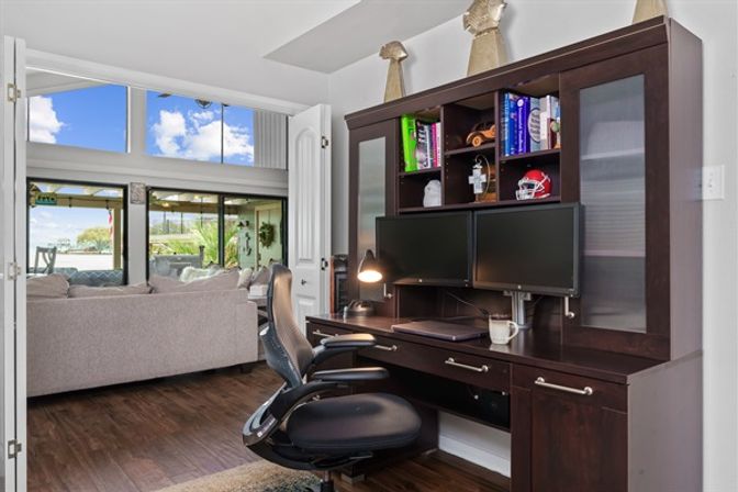 Bright home office with dark wood desk and dual monitors, ergonomic chair and bookshelves, opening to a living room and sliding doors with a waterfront patio view.