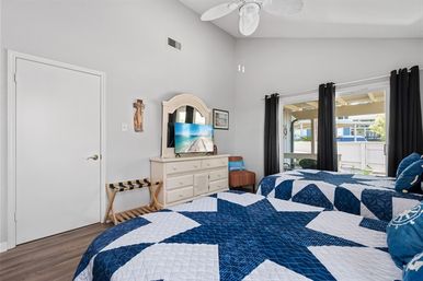 Bright coastal-themed bedroom with two twin beds in blue star quilts, white dresser with mirror and flat-screen TV, ceiling fan, and sliding glass door to a patio.