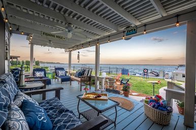 Cozy waterfront covered porch at sunset with blue cushioned seating, string lights, coffee table with snacks, grassy yard, dock and boats on the water.