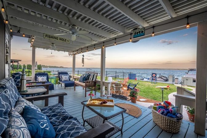 Cozy waterfront covered porch at sunset with blue cushioned seating, string lights, coffee table with snacks, grassy yard, dock and boats on the water.