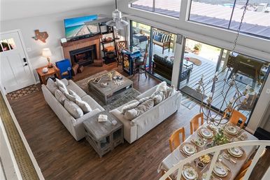 Aerial view of an open-plan living and dining area with a light-gray sectional sofa, wood coffee table and fireplace with TV, hardwood floors, chandelier-lit dining table set for six, and large sliding glass doors opening to a deck with outdoor seating and grill.