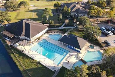 Aerial view of a sunlit suburban community pool complex featuring a large rectangular lap pool, two smaller recreational pools, poolside lounge chairs, covered pavilion and clubhouse beside landscaped lawns, parking lot and pond.