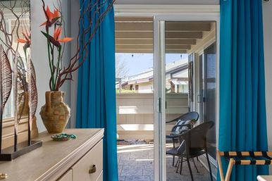 Interior view through sliding glass doors to a small covered patio with two wicker chairs, bright turquoise curtains framing the doorway, and a decorative vase with bird-of-paradise flowers on a dresser.