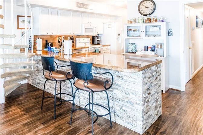 Bright coastal-style kitchen with stone-faced breakfast bar and polished countertop, two metal swivel bar stools, white cabinets, spiral staircase and warm wood-look flooring