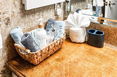 Rustic bathroom vanity with tan countertop, woven basket of rolled blue and gray towels, lotion pump and cotton swabs, folded fan towel and dark toothbrush holder by a mirror.