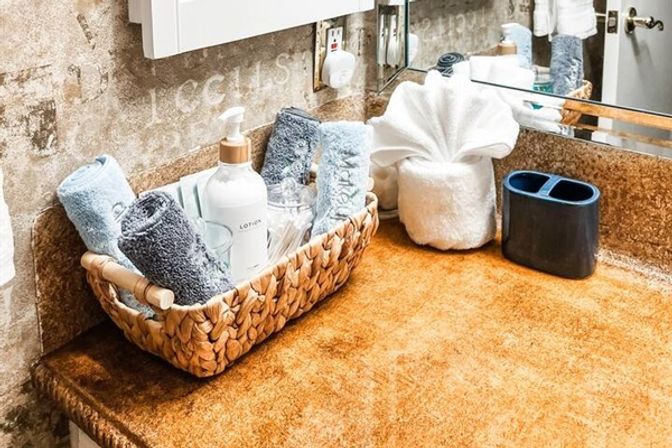 Rustic bathroom vanity with tan countertop, woven basket of rolled blue and gray towels, lotion pump and cotton swabs, folded fan towel and dark toothbrush holder by a mirror.