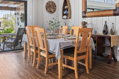 Sunlit coastal dining room with a wooden table set for six, light-wood chairs, fringed tablecloth and eucalyptus centerpiece, sliding glass door opening to a porch with a rocking chair and tropical greenery.