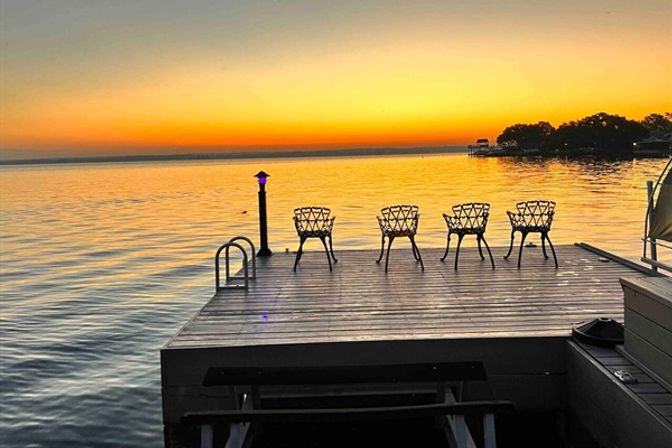 Vibrant orange sunset over a calm lake, a wooden dock with four decorative metal chairs and a lit post, silhouetted shoreline in the distance.