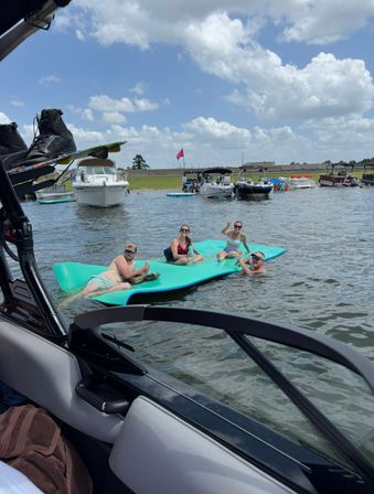 Four adults lounging on a bright turquoise floating mat near anchored boats on a sunny lake, blue sky with fluffy clouds and a grassy shoreline in the background.