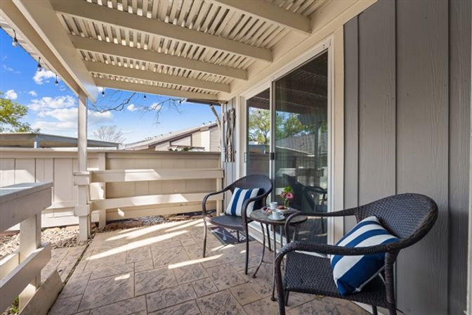 Covered residential patio with two black wicker chairs with blue-and-white striped cushions, small round table holding a teacup and flowers, sliding glass door, beige privacy railing and stamped concrete floor under a pergola-style roof with string lights.