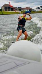 Wakeboarder smiling and flexing while riding a wake on a suburban lake, holding a red tow rope and wearing a life vest and patterned shorts with waterfront homes in the background