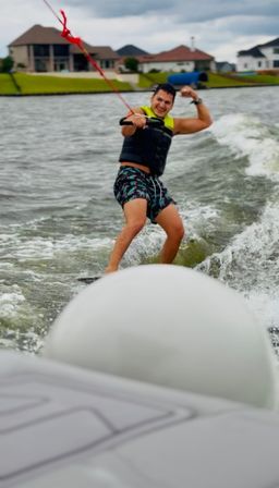 Wakeboarder smiling and flexing while riding a wake on a suburban lake, holding a red tow rope and wearing a life vest and patterned shorts with waterfront homes in the background