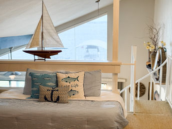 Coastal loft bedroom with nautical decor: model sailboat on a railing by a large window, striped bedding with fish-themed and anchor pillows, woven rug and white staircase.