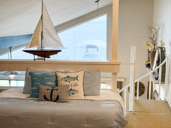 Coastal loft bedroom with nautical decor: model sailboat on a railing by a large window, striped bedding with fish-themed and anchor pillows, woven rug and white staircase.