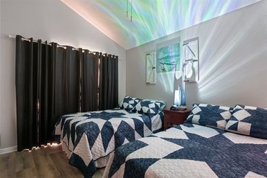 Cozy coastal-inspired bedroom with two twin beds in navy-and-white geometric quilts, a small wooden nightstand with a glowing lamp casting colorful light patterns on the vaulted ceiling, dark blackout curtains and wood-look flooring.