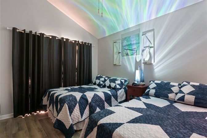 Cozy coastal-inspired bedroom with two twin beds in navy-and-white geometric quilts, a small wooden nightstand with a glowing lamp casting colorful light patterns on the vaulted ceiling, dark blackout curtains and wood-look flooring.