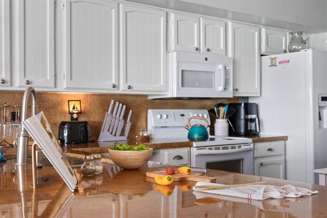 Bright white kitchen with glossy brown granite island, white cabinets, microwave and stove, teal kettle on range, knife block, toaster, wooden salad bowl and cutting board with peppers.