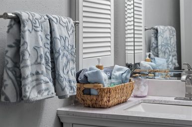 Spa-like bathroom vanity with light blue patterned towels on a towel bar, a woven basket of rolled towels and a soap pump on a marble countertop, and a mirror reflecting the arrangement.