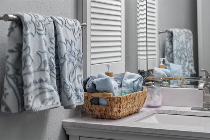 Spa-like bathroom vanity with light blue patterned towels on a towel bar, a woven basket of rolled towels and a soap pump on a marble countertop, and a mirror reflecting the arrangement.