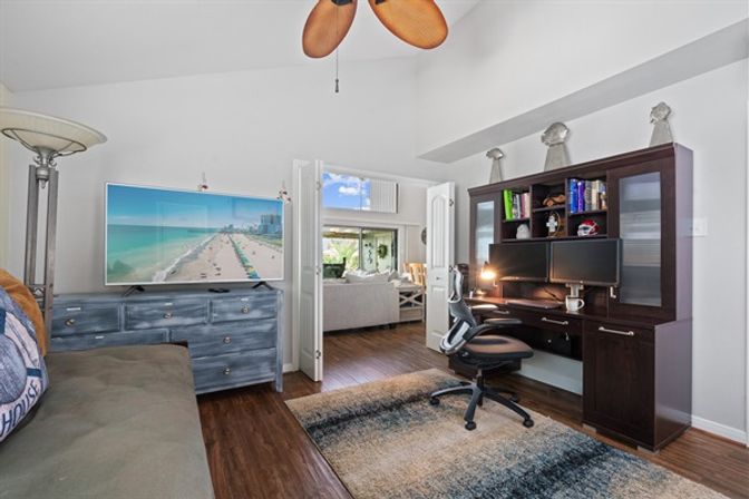 Sunny coastal-themed home office with dark wood desk and dual monitors, ergonomic chair, TV showing a sandy beach on a blue dresser, ceiling fan, rug, and open French doors into a bright living room