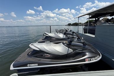 Three silver-gray Yamaha jet skis lined up on a lakeside dock under a sunny blue sky with puffy clouds