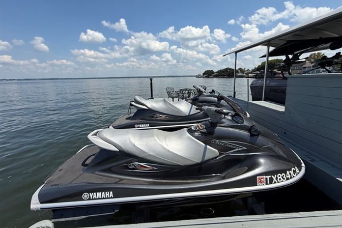 Three silver-gray Yamaha jet skis lined up on a lakeside dock under a sunny blue sky with puffy clouds