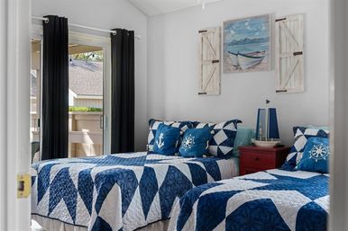 Cheerful coastal guest bedroom with two twin beds in blue-and-white nautical quilts, anchor and ship‑wheel pillows, red nightstand with sailboat lamp, boat painting with shutter decor, and sliding glass door to a sunlit balcony.