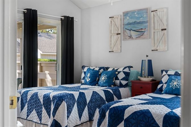Cheerful coastal guest bedroom with two twin beds in blue-and-white nautical quilts, anchor and ship‑wheel pillows, red nightstand with sailboat lamp, boat painting with shutter decor, and sliding glass door to a sunlit balcony.