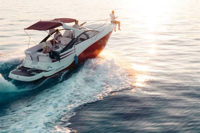 Sunset motorboat cruise on calm open sea — red-and-white cruiser slicing through water with passengers lounging and one person perched on the bow, sun sparkling on the wake.