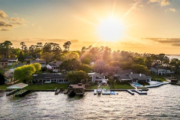 Golden sunset over a waterfront neighborhood — tree-lined shoreline with lakeside homes, private docks, boats and calm water reflecting warm light.