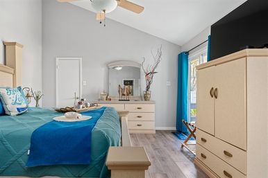 Coastal-inspired bedroom with teal quilt, beige wood dresser and armoire, blue curtains, sliding glass door and ceiling fan.