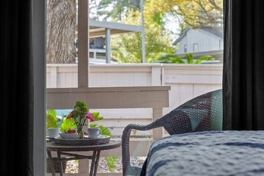 View through a bedroom sliding glass door to a cozy sunlit patio with a round table holding coffee cups and pink flowers, a wicker chair, and a white wooden fence