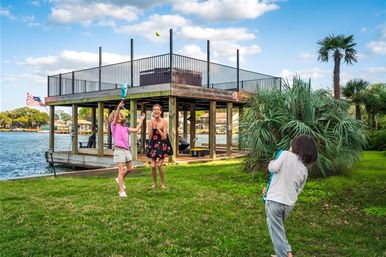 Three people playing catch with a ball on a grassy lakeside lawn by a raised wooden dock and palm trees under a sunny blue sky