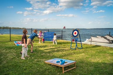 Kids playing lawn games on a lakeside waterfront lawn with cornhole, giant Connect Four and a target toss near a dock and jet skis under a blue sky