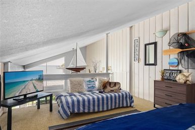Coastal loft bedroom with sloped ceiling and large water view, nautical décor with sailboat model, oars and 'Relax' sign, blue-and-white striped daybed, wooden dresser and TV showing a sunny beach.