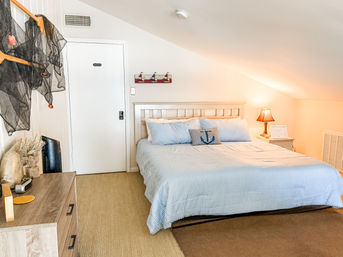 Cozy sunlit coastal attic bedroom with sloped ceiling, king bed dressed in blue-and-white striped bedding, anchor throw pillow, wooden headboard, bedside lamp and nautical accents.